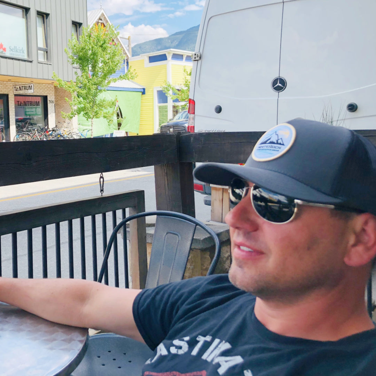 Man wearing black outdoors hat and sunglasses sitting at metal table in outdoor cafe with colorful buildings and white van in background