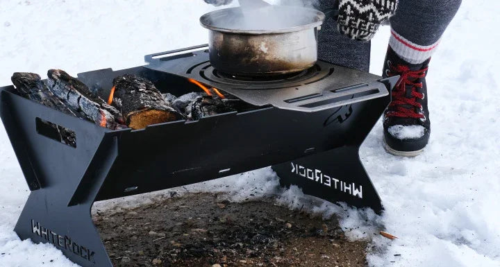Black Whitrock portable fire pit with burning logs and steaming pot in snow, person wearing winter boots nearby