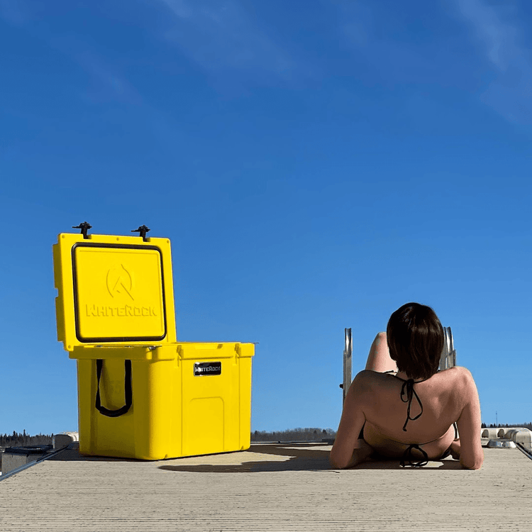 Woman sunbathing on dock next to bright yellow WinterRock cooler under clear blue sky