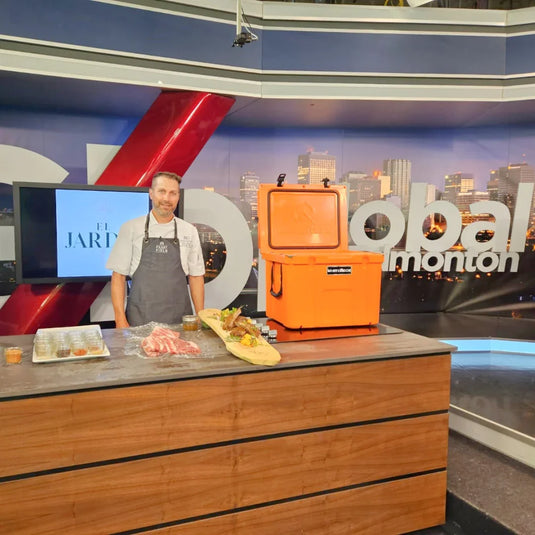 Chef in apron standing behind kitchen counter with meat cuts, prepared dishes, and orange cooler in TV studio