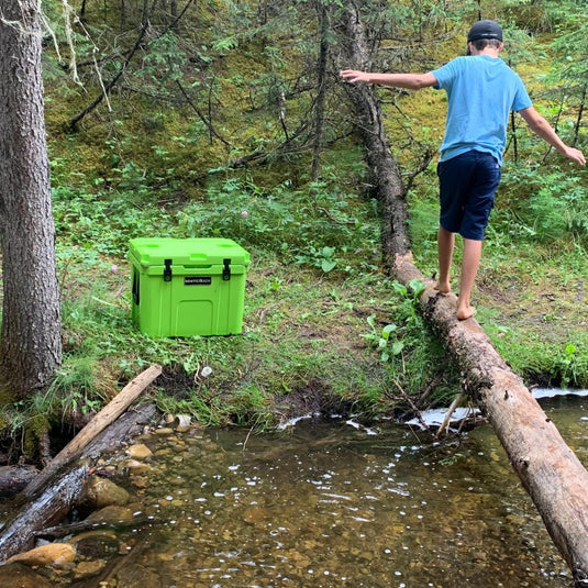 bright green WhiteRock cooler on grass by creek with boy barefoot balancing on log in forest