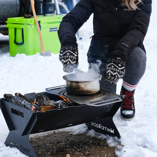 Person wearing mittens stirring pot on White Rock portable camping fire pit in snowy outdoor setting