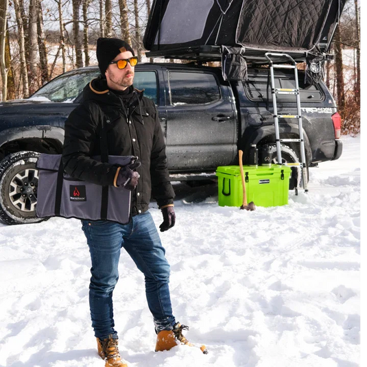Black carrying bag for fire pit held by man in winter jacket, standing in snowy campsite with off-road truck and rooftop tent
