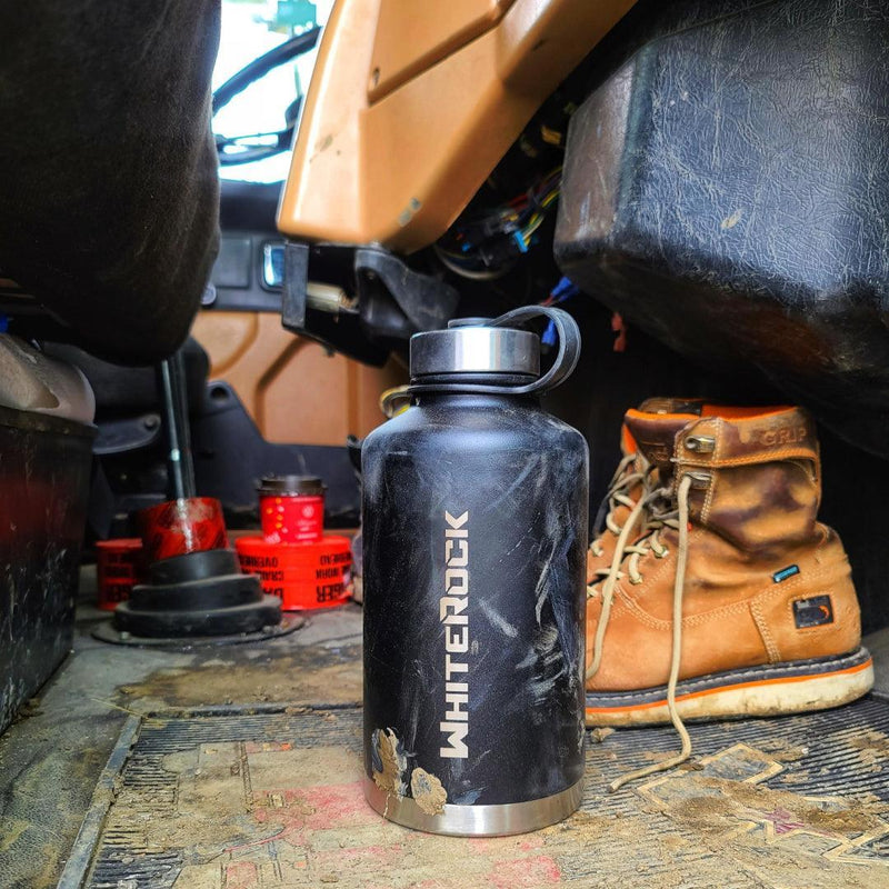 Load image into Gallery viewer, Black WhitRock water bottle with mud splatters inside a dirty vehicle cabin, next to dusty brown work boots
