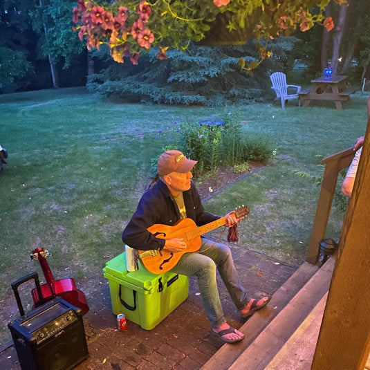 Man in cap playing vintage wooden guitar outdoors on green cooler near red guitar and amplifier