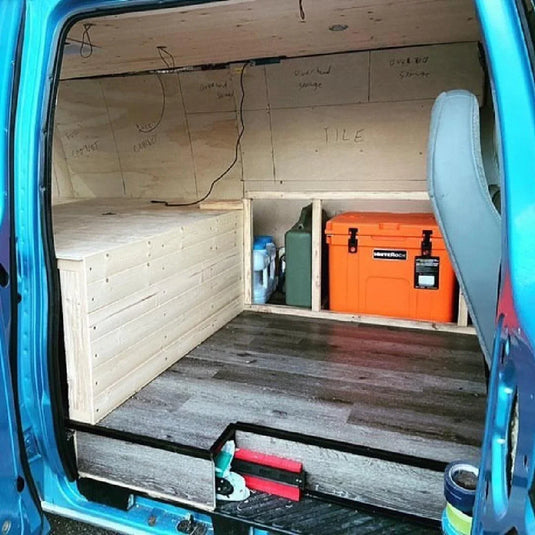 Blue van interior with custom wooden storage platform, gray vinyl flooring, and orange storage cooler