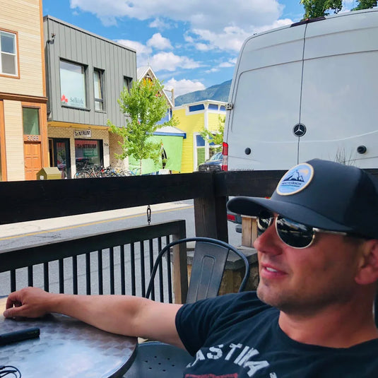 Man wearing gray Winterduck cap and sunglasses sitting at outdoor metal table in small town setting