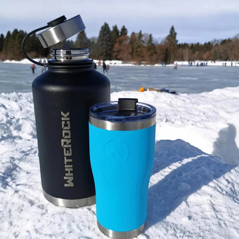Load image into Gallery viewer, Black WhiteRock insulated bottle and blue travel tumbler on snowy surface near frozen lake with people ice skating

